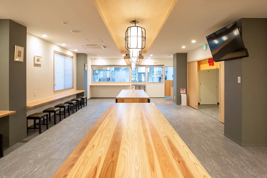 広々とした木目調のシェアスペースの全景。 空間の奥に向かって、長い木製テーブルが2台並び、左側の壁沿いにはカウンター席とスツールが見えます。