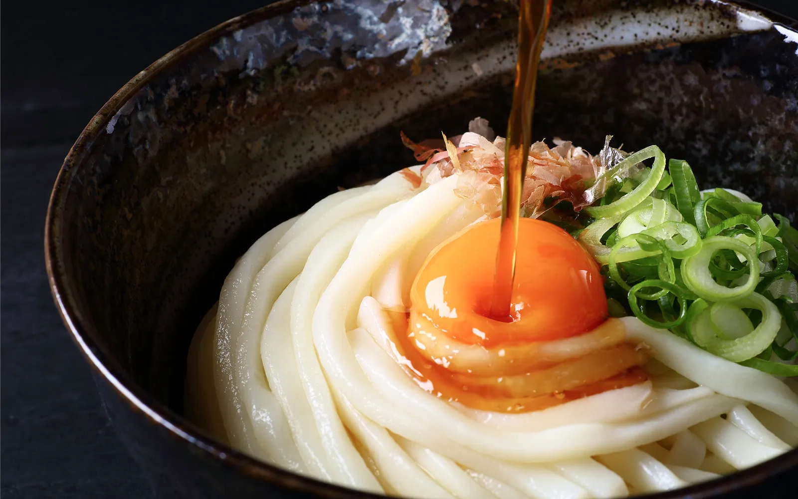 釜揚げうどんに、つやつやとした卵黄と刻みネギ、鰹節がトッピングされ、上から特製の醤油がとろりと注がれています。