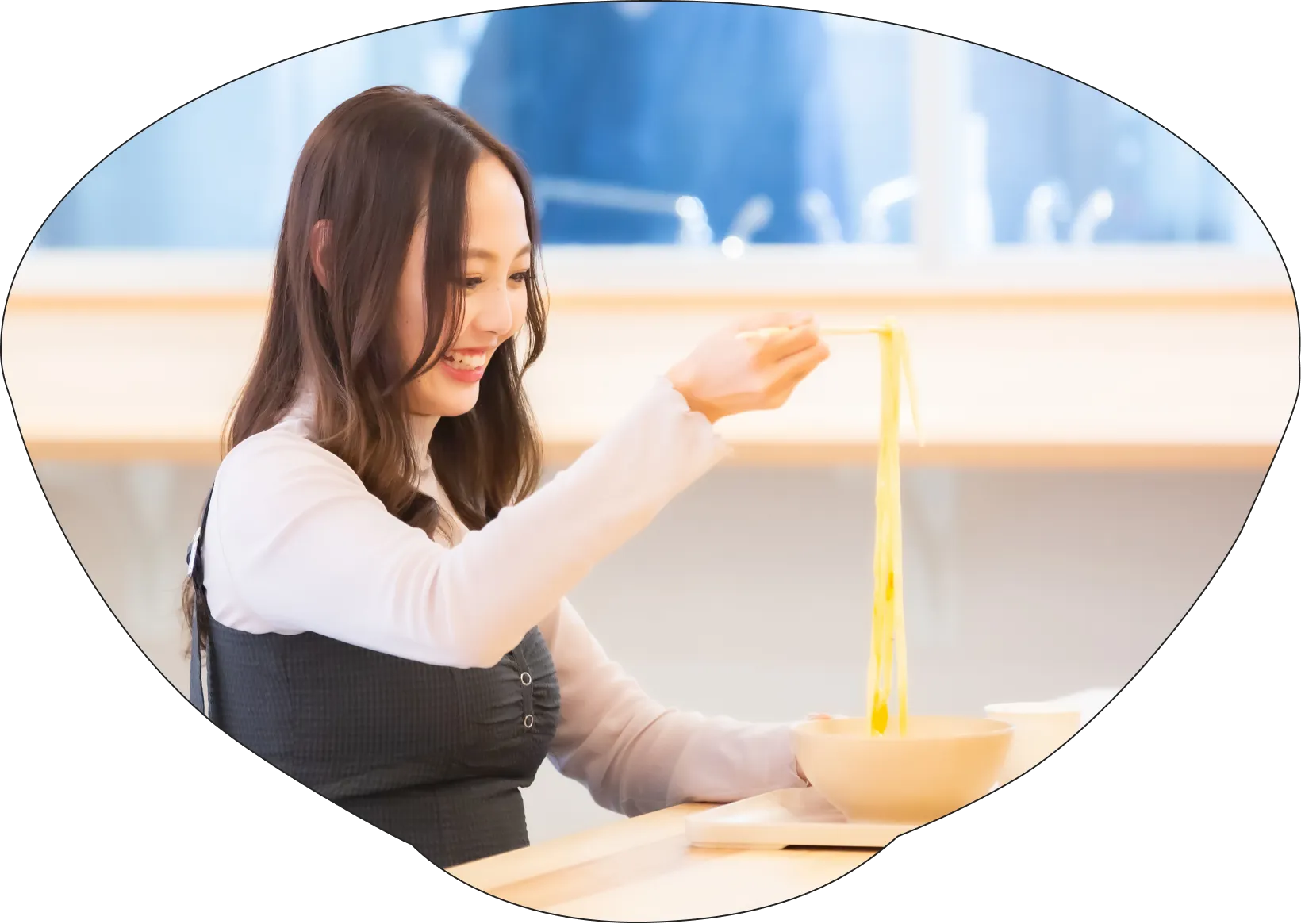 明るい店内で、笑顔の女性が箸で釜揚げ風うどんを高く持ち上げている様子。