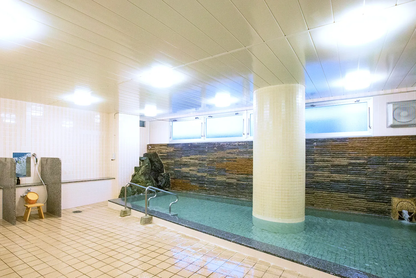 明るい照明とベージュのタイルが清潔な印象を与える、内湯。右奥に細長い浴槽があり、湯船の奥の壁は茶色と黒の石積み、左手前には洗い場と木製の椅子と桶が見える。