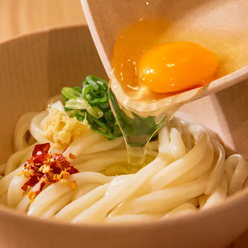 艶やかなうどんの上に、器から生卵の卵黄と白身が流れ落ちている様子。薬味として、ネギ、おろし生姜、唐辛子が添えられている写真の1枚目