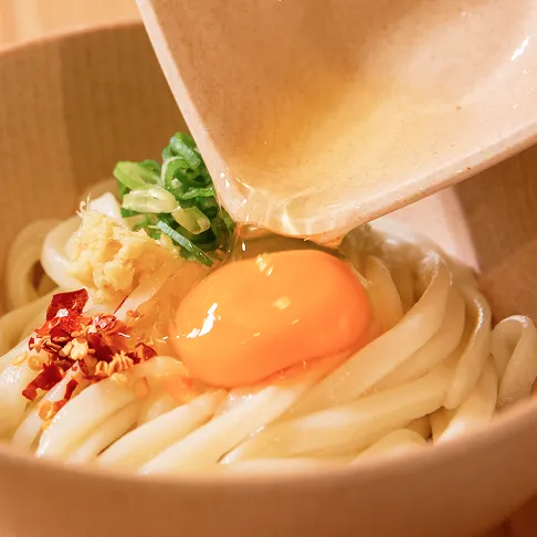 艶やかなうどんの上に、器から生卵の卵黄と白身が流れ落ちている様子。薬味として、ネギ、おろし生姜、唐辛子が添えられている写真の2枚目
