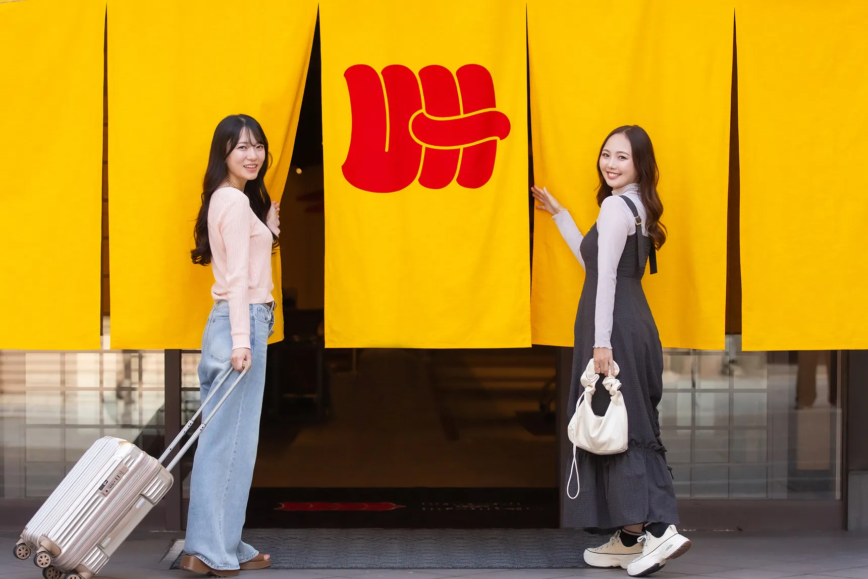 鮮やかな黄色の暖簾の前に立つ二人の若い女性。左の女性はキャリーケースを引き、右の女性は笑顔で暖簾に手を添えている写真の1枚目