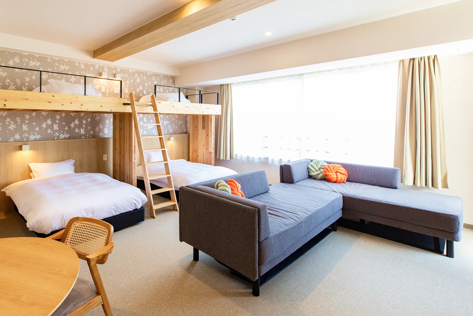 ロフトベッドと大きなL字型ソファが置かれた、カーペット敷きの客室。左奥に木製のロフトベッドがあり、下に2台、上にも2台のベッドが設置されている。手前中央にはグレーのL字型ソファがあり、オレンジと緑の丸いクッションがアクセントになっている。