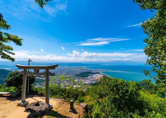 鳥居と石灯籠がある山頂の展望台から、眼下に広がる街並みと青い海、そして遠くの山々を一望できる景色。空は雲が少なく、晴れ渡っている。