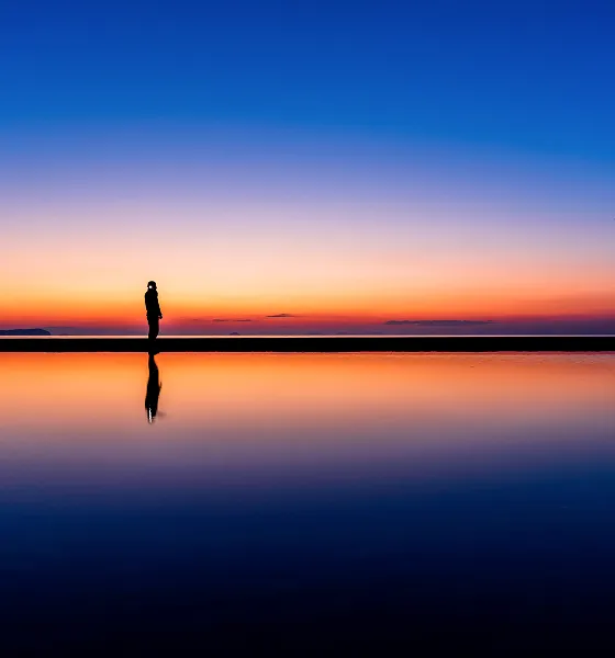 夕焼けに染まる空と海を背景に、水面に立つ人物のシルエット。空の色は上から紺色、紫色、オレンジ色へとグラデーションになり、静かな水面にはそれらが鏡のように映り込んでいる。