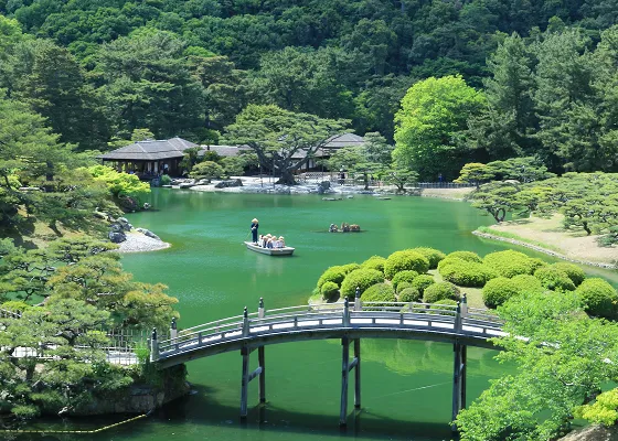 広大な日本庭園の池。緑色に澄んだ水面には、舟に乗った人が数人いる。池の中央には小さな島があり、その周りを曲がりくねった橋が囲んでいる。
