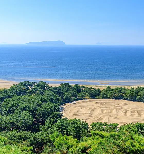 展望台から見下ろす、砂浜に描かれた巨大な「寛永通宝」の砂絵。その手前には緑豊かな松林が広がり、奥には青い海と砂浜、そして遠くの島々が見える。