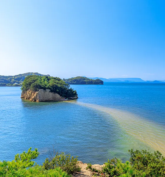 青い海に浮かぶ、緑に覆われた岩の島々。手前には小石と岩が混じったような色の道が海中に伸びており、潮の満ち引きによって現れる「天使の散歩道」のような神秘的な景色。